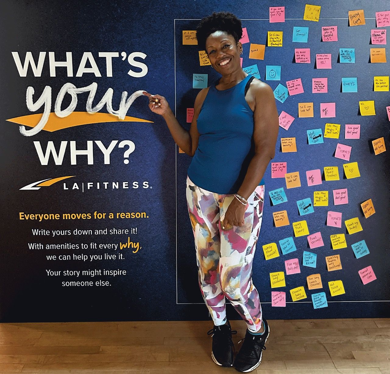 A woman in workout attire smiles next to the "What's Your Why?" wall covered in colorful sticky notes at the LA Fitness community activation.
