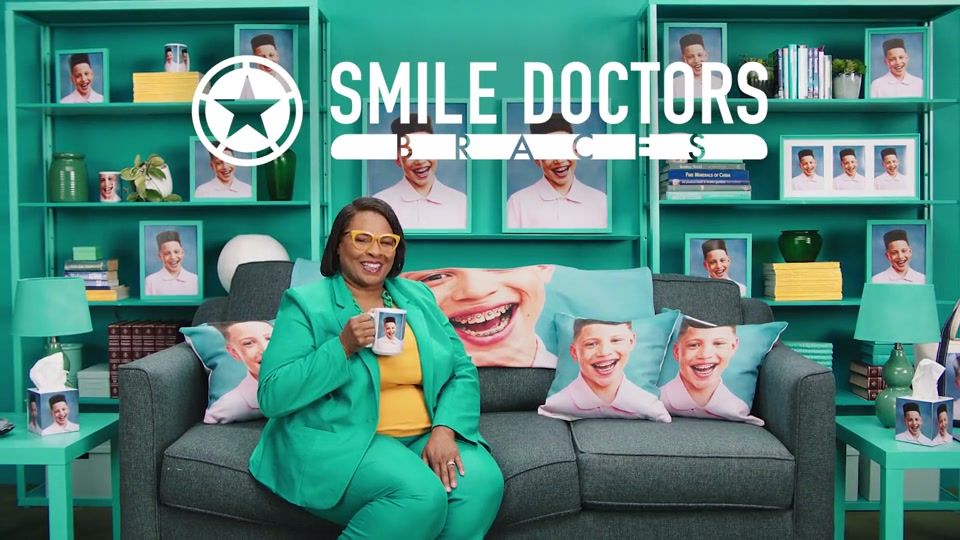 A proud mom sits on a couch layered with throw pillows featuring her son's braced smile, shelves of matching framed photos behind her, under the Smile Doctors Braces logo.