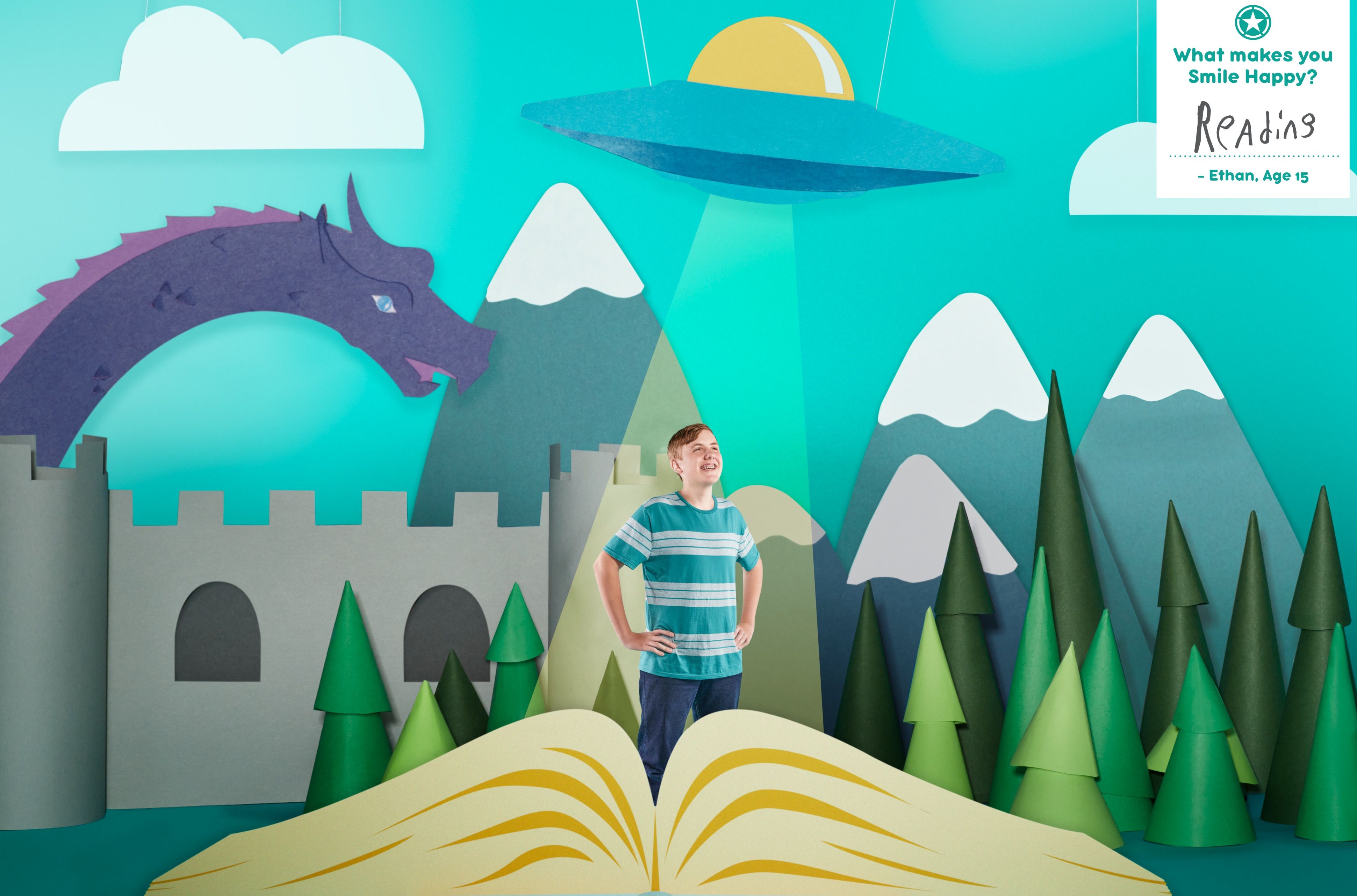 Campaign photo of Ethan, age 15, standing in a handcrafted paper-art world with a castle, dragon, and UFO, captioned "What makes you Smile Happy? Reading."