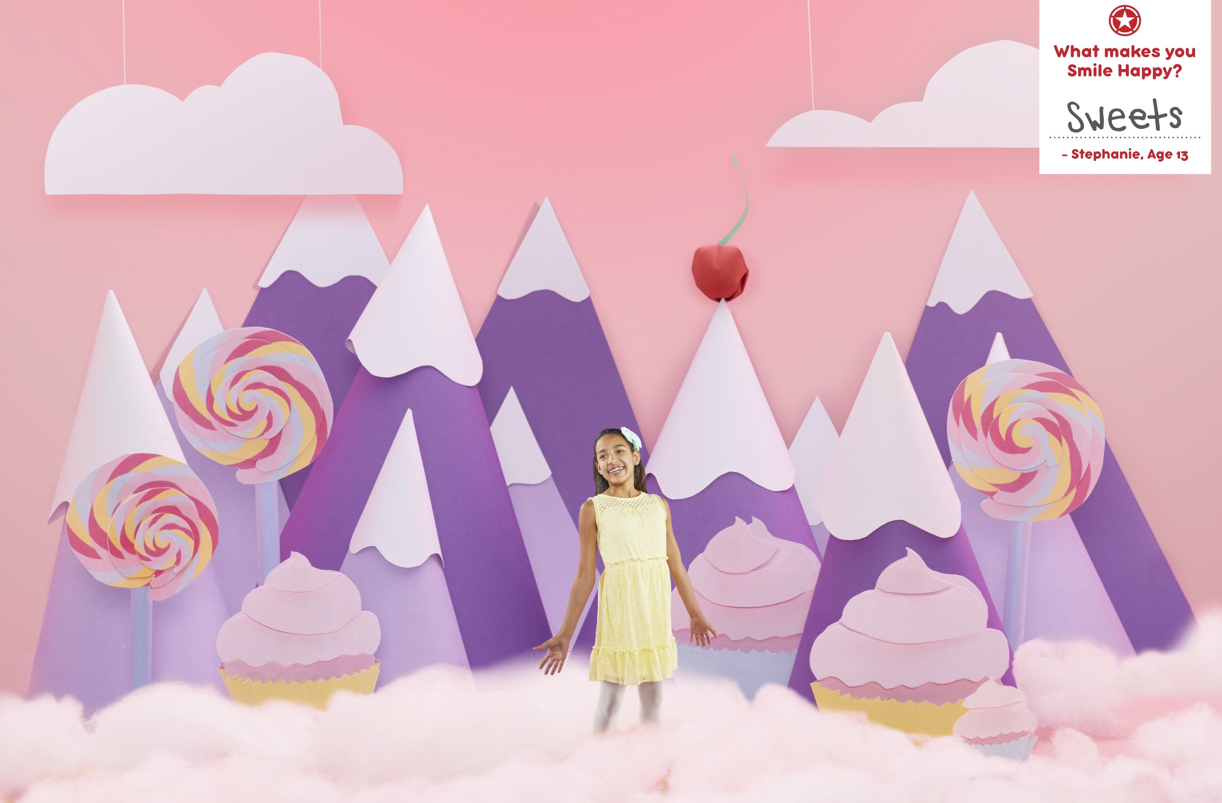 Campaign photo of Stephanie, age 13, standing in a paper-art candy land of giant lollipops and cupcakes, captioned "What makes you Smile Happy? Sweets."