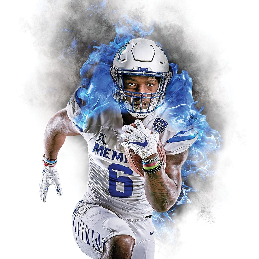 Close-up portrait of Memphis Tigers running back #6 charging forward with a football tucked tight, intense blue fire and smoke erupting around him.